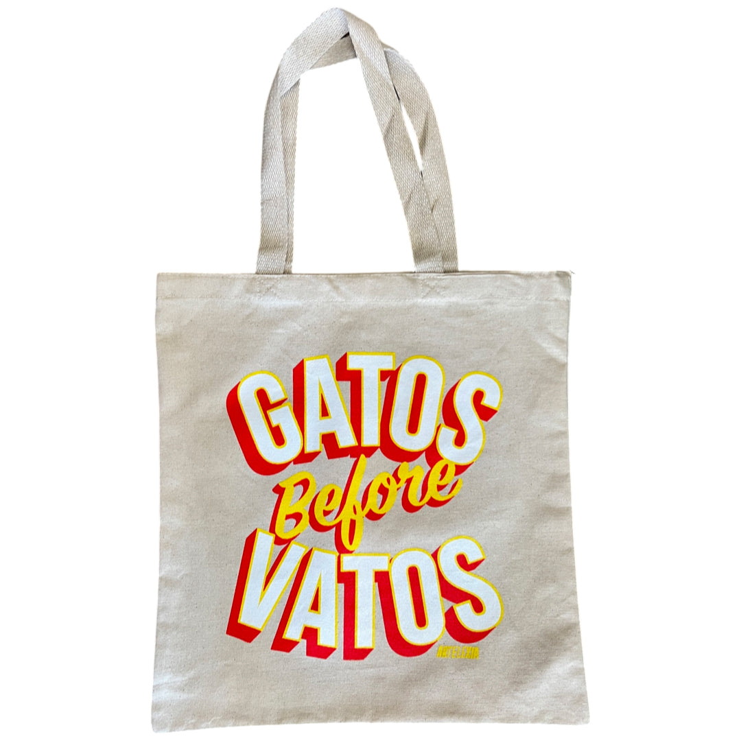 Natural colored canvas tote bag featuring the phrase "Gatos Before Vatos" in white letters with yellow outline and red drop shadow
