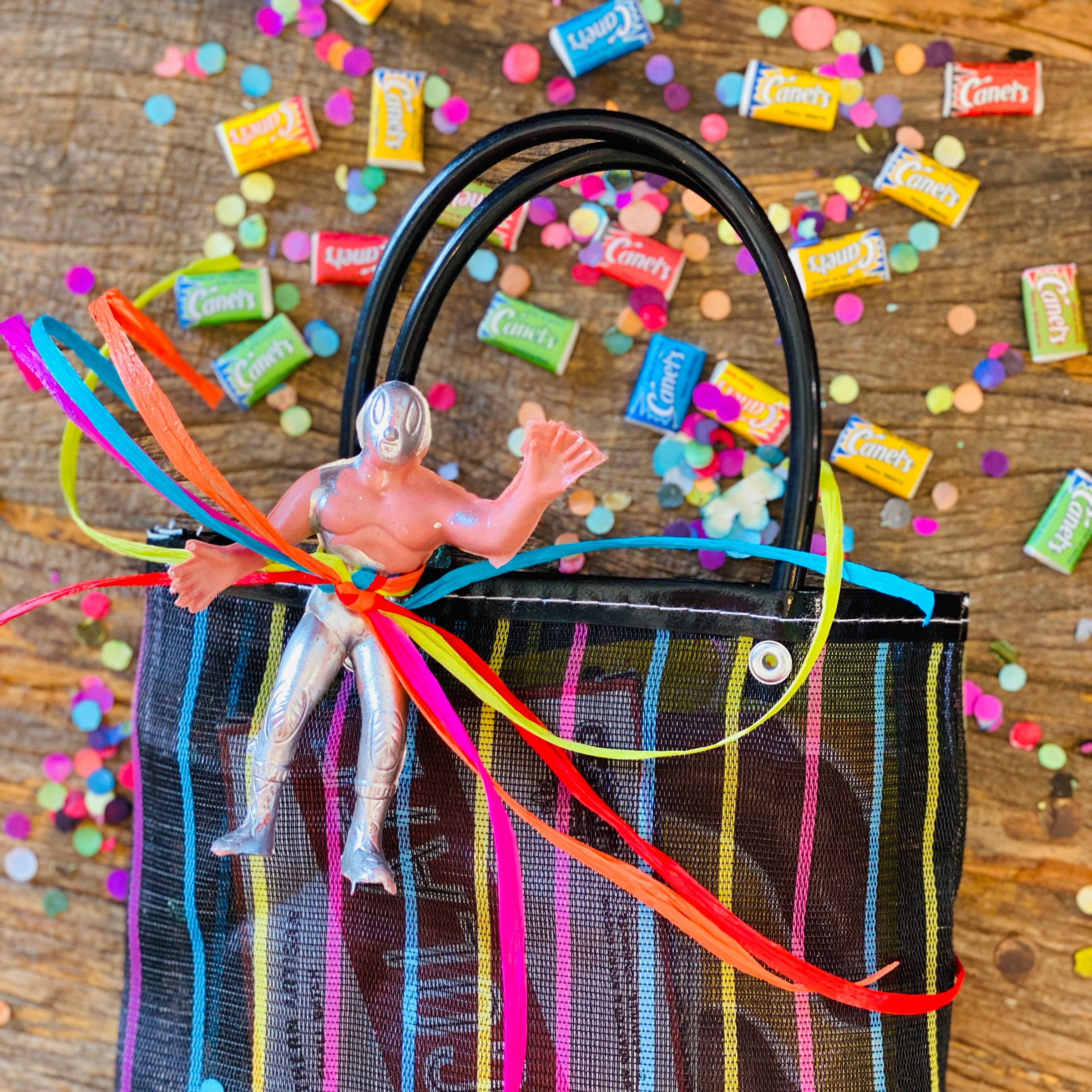Top view of goodie bag with luchador figure tied to bag strap with colorful bow. Goodie bag on wooden table, sprinkled with confetti and gum.
