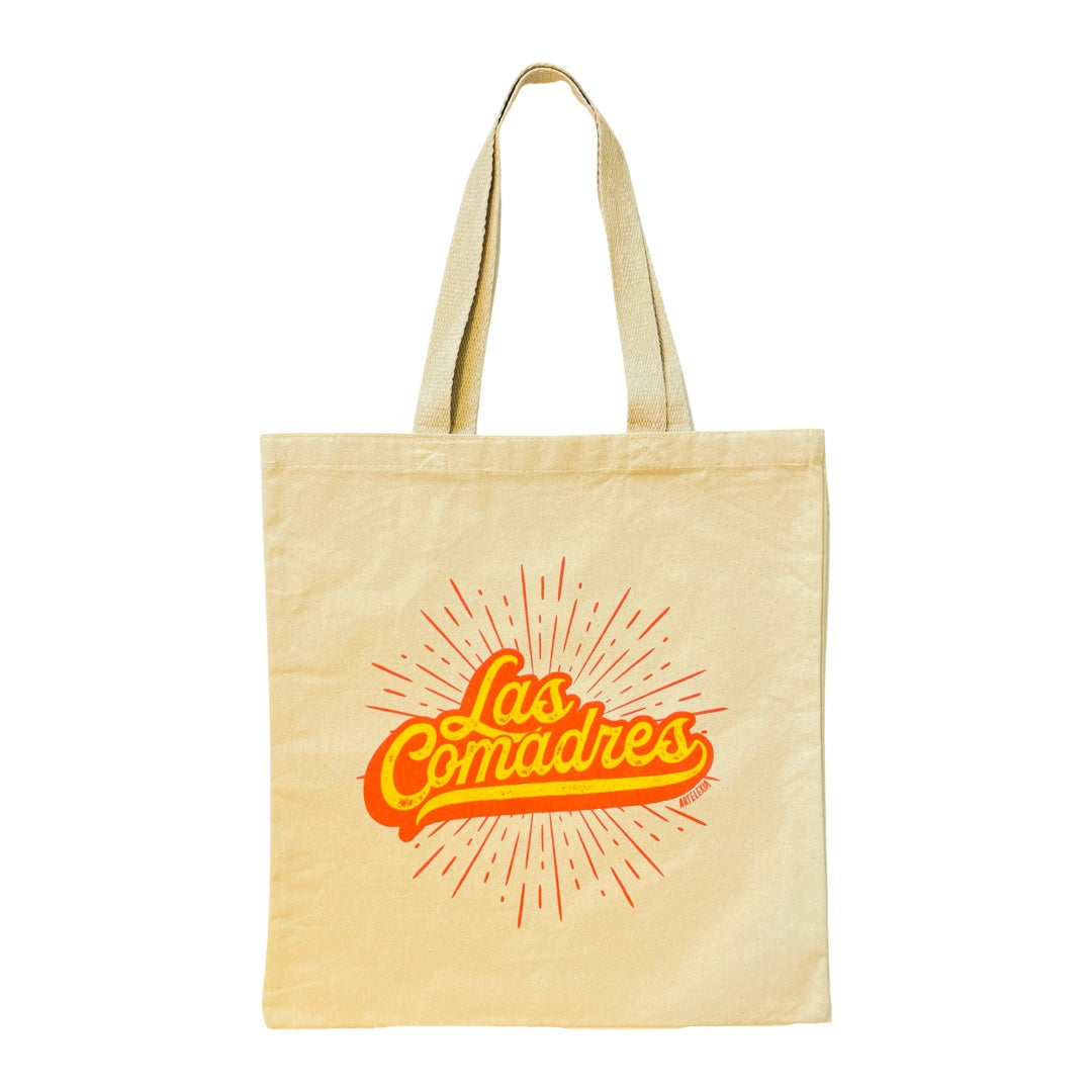 Natural canvas tote bag with the phrase Las Comadres in orange and yellow lettering with orange rays in the background.