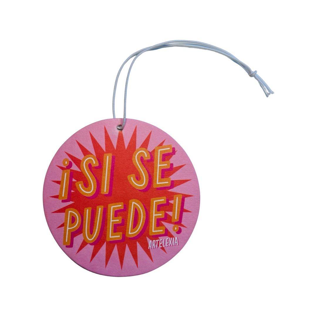 PInk round air freshener with the phrase Si Se Puede in orange and white lettering with an orange burst inthe background.