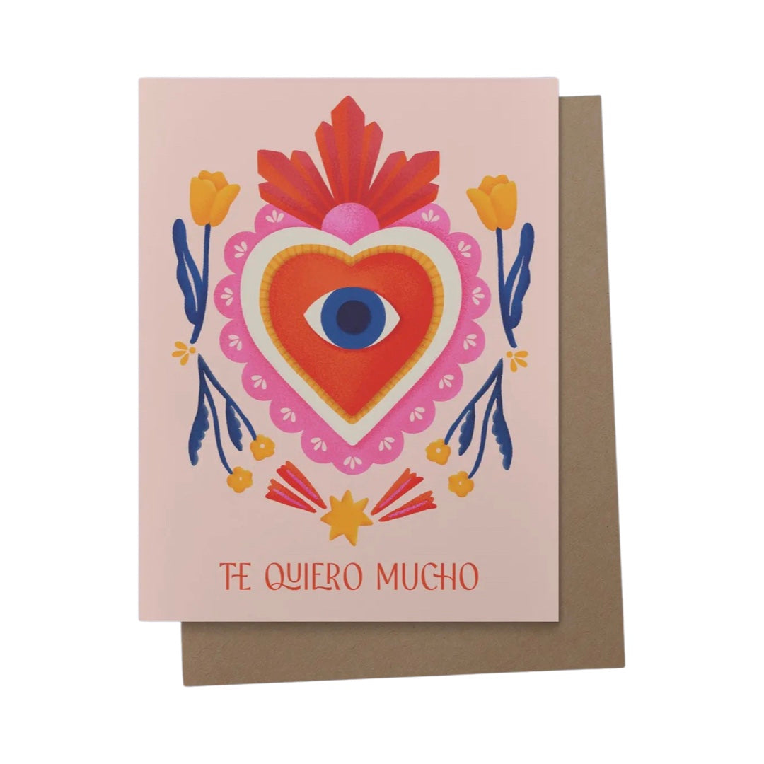 Pink card with a pink and red milagro heart design featuring an eye in the center and orange flowers. Includes the phrase Te Quiero mucho in red lettering.
