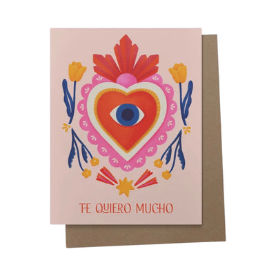 Pink card with a pink and red milagro heart design featuring an eye in the center and orange flowers. Includes the phrase Te Quiero mucho in red lettering.