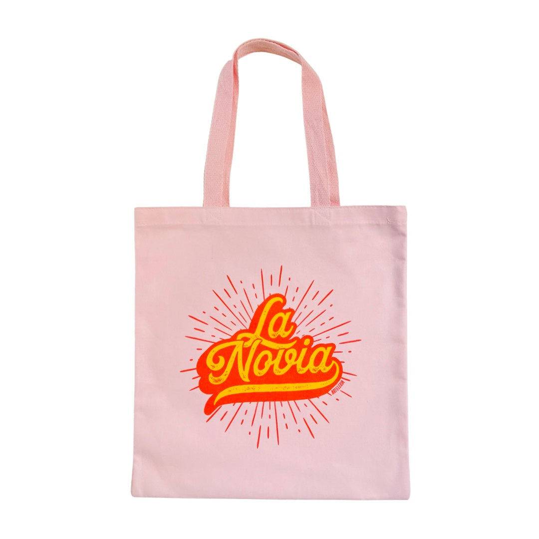 Pink canvas tote bag with the phrase La Novia, The Bride, in yellow lettering outlined in red with a red burst in the background