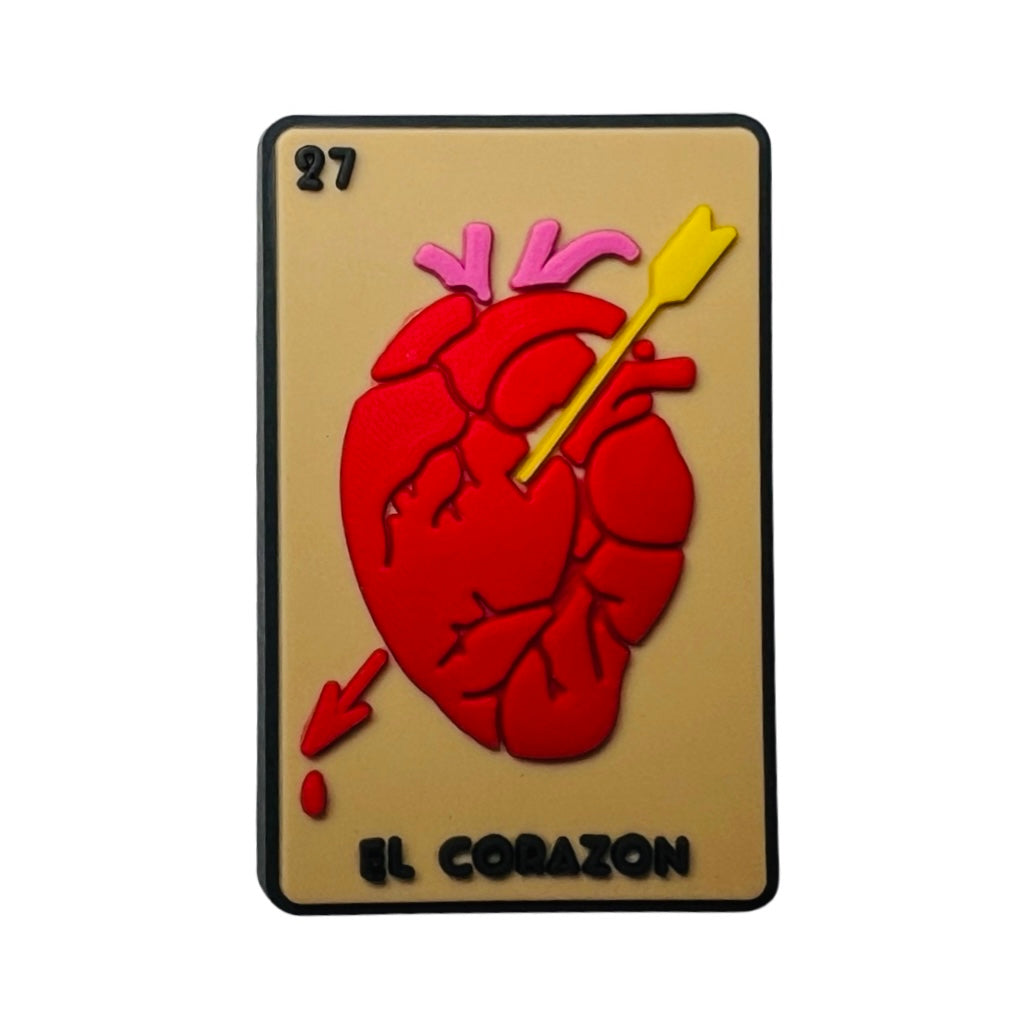 Beige rubber chancla, croc, charm with a red heart and arrow and the phrase El Corazon in black lettering. 