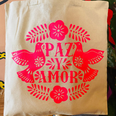 Close up view of natural colored tote bag with bright pink design of two doves and the words "Paz y amor" in the center.