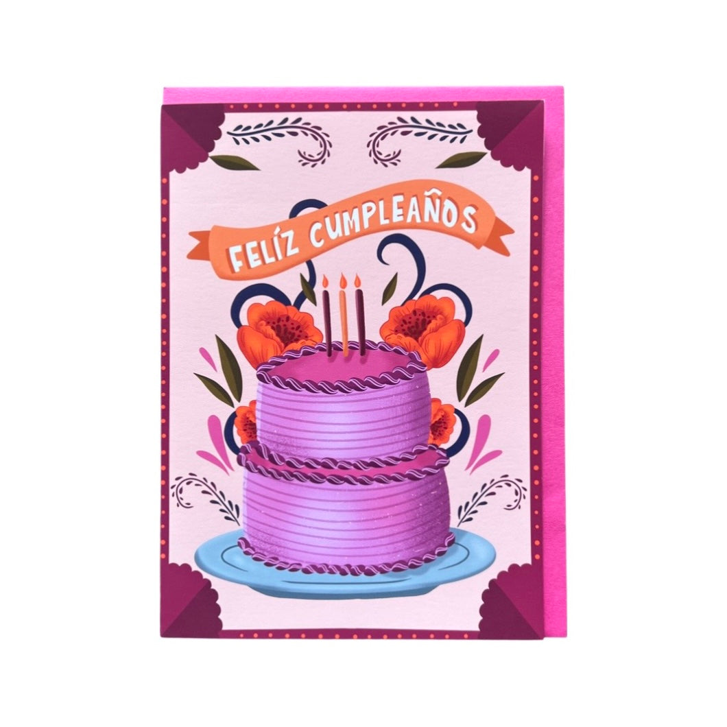 Pink and purple greeting card with a purple two tier cake and orange flowers featuring an orange banner above the cake with the phrase Feliz Cumpleanos in white lettering.
