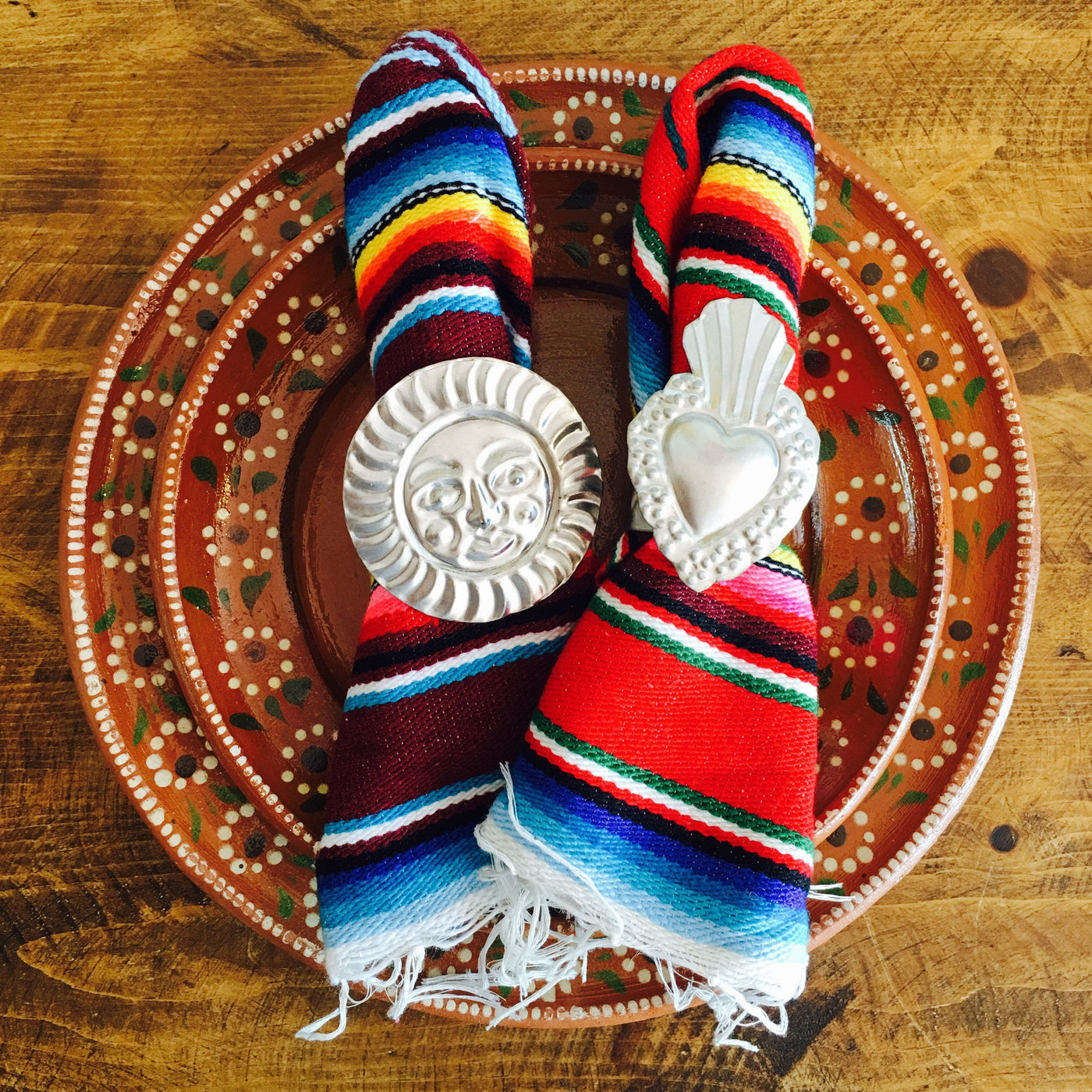 Tin napkin rings pictured with serape napkins on top of red floral clay plate. Napkin ring designs featured: el sol (sun) and sacred heart.  