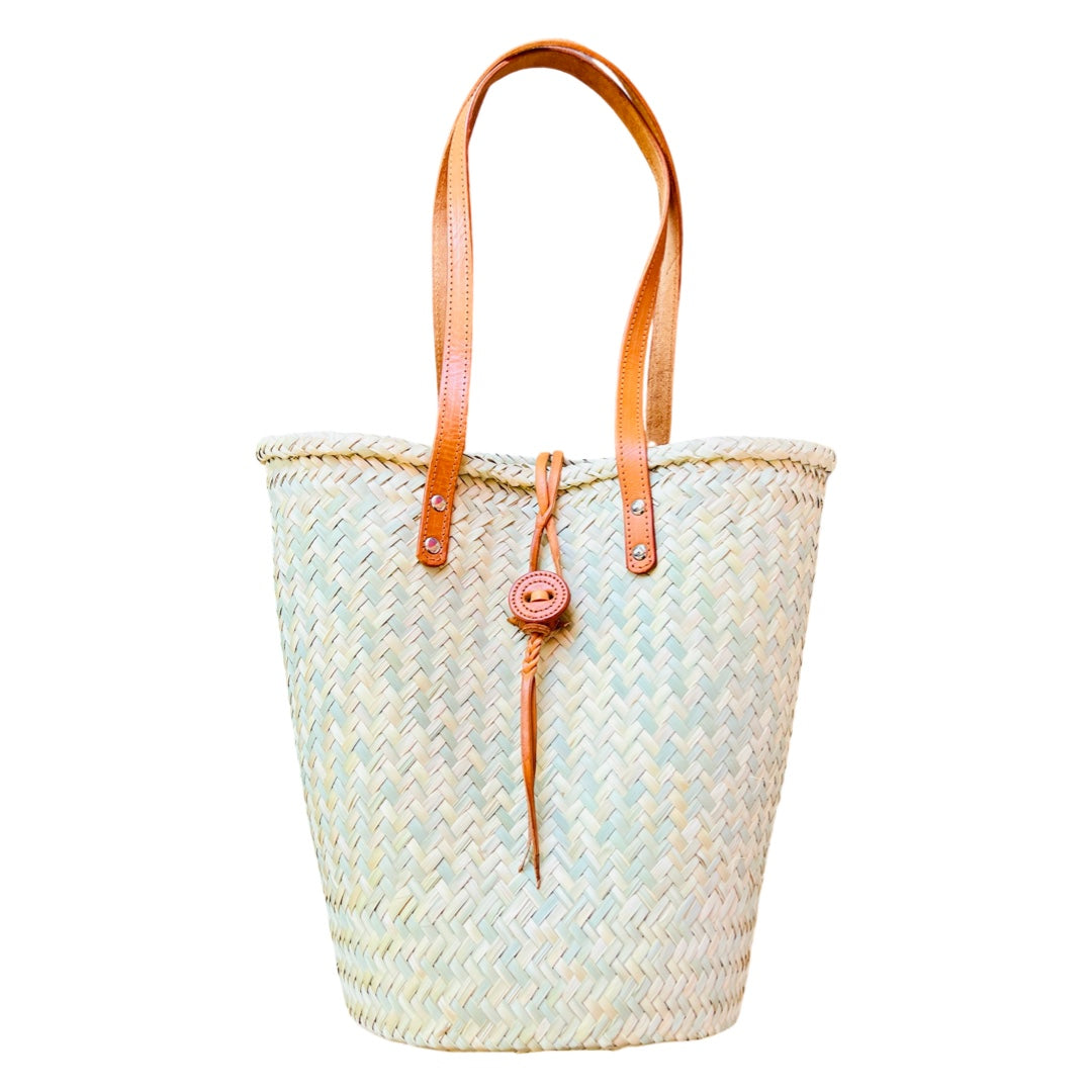 Single palm leaf woven bag that tapers from the top to the bottom and features two leather straps and bag closure.