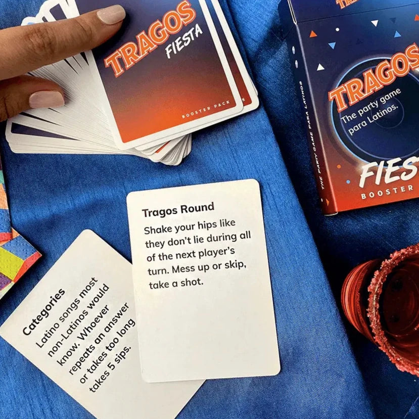 Tragos Fiesta Booster Pack cards displayed on a blue cloth, featuring bilingual prompts such as "Shake your hips like they don’t lie" and "Latino songs most non-Latinos would know." The scene includes a hand holding cards, a colorful game box, and a festive drink with a red rim.