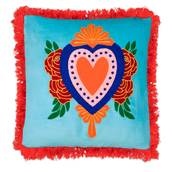 Square teal pillow with a central embroidered pink, blue and orange heart, floral accents, and bright orange fringe trim.