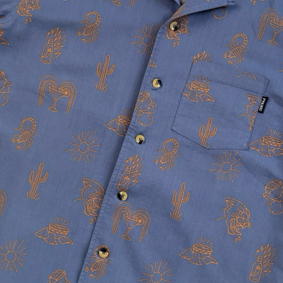 Close up view of a Blue short-sleeve button-down shirt with allover brown graphic prints including cacti, suns, roosters, and southwest-inspired designs.