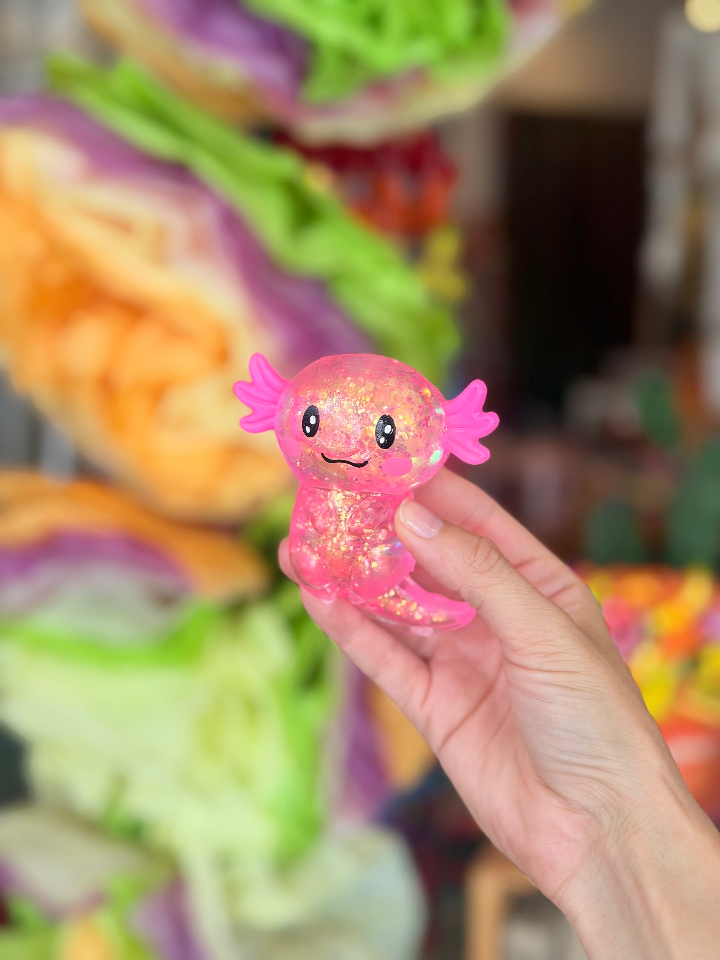 A hand holding a small, glittery pink axolotl squishy sensory toy with a cute face, set against a colorful, blurred background.