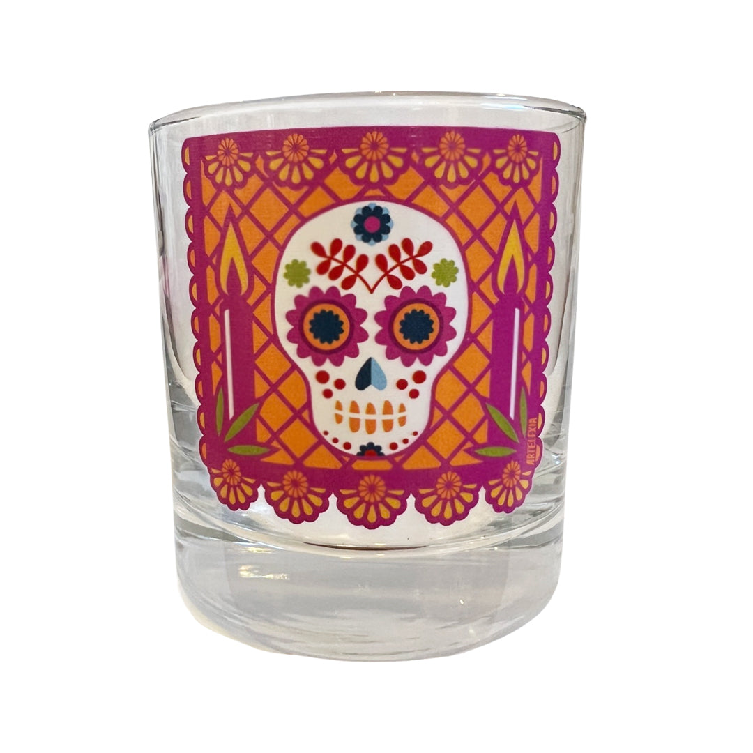 Photo of side view of cocktail glass featuring a berry colored papel picado with a sugar skull in the center & an orange background