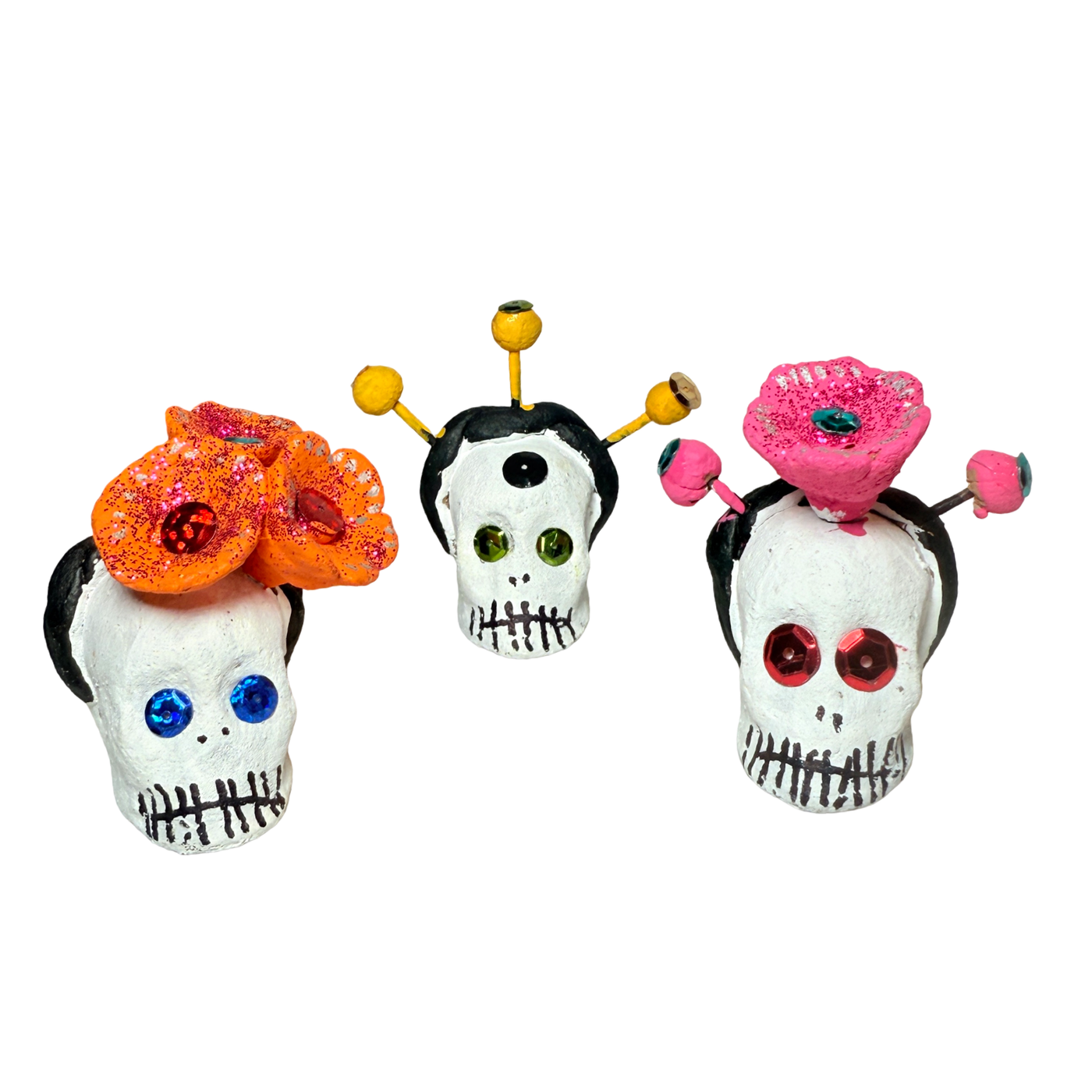 Three handcrafted Día de Muertos calavera skulls with jeweled eyes and colorful accents—one with orange glitter marigolds, one with yellow pods, and one with a pink flower crown.