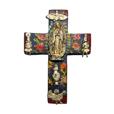 A hand-painted wooden cross with colorful floral designs, featuring a gold-tone Virgen de Guadalupe at the center. The cross is decorated with various milagro charms, including a heart, rooster, and hand, symbolizing faith and blessings