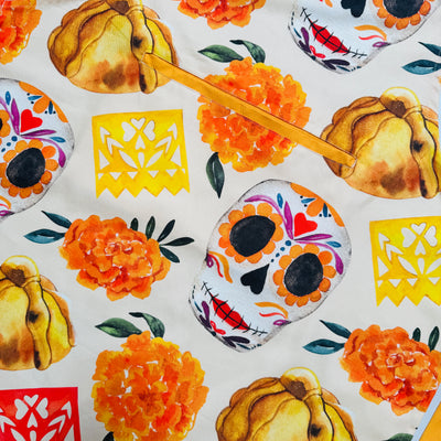 close up view of a Cream apron printed with Día de Muertos motifs—sugar skulls, orange marigolds, pan de muerto, and yellow, orange, and purple papel picado.