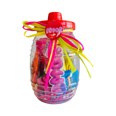 A clear candy jar decorated with vibrant ribbons and filled with colorful confetti, assorted Mexican candies, and a pink and white pin-back button featuring a red background and the word AMOR in pink bubble lettering. The jar also includes sticker of the word AMOR in pink bubble lettering