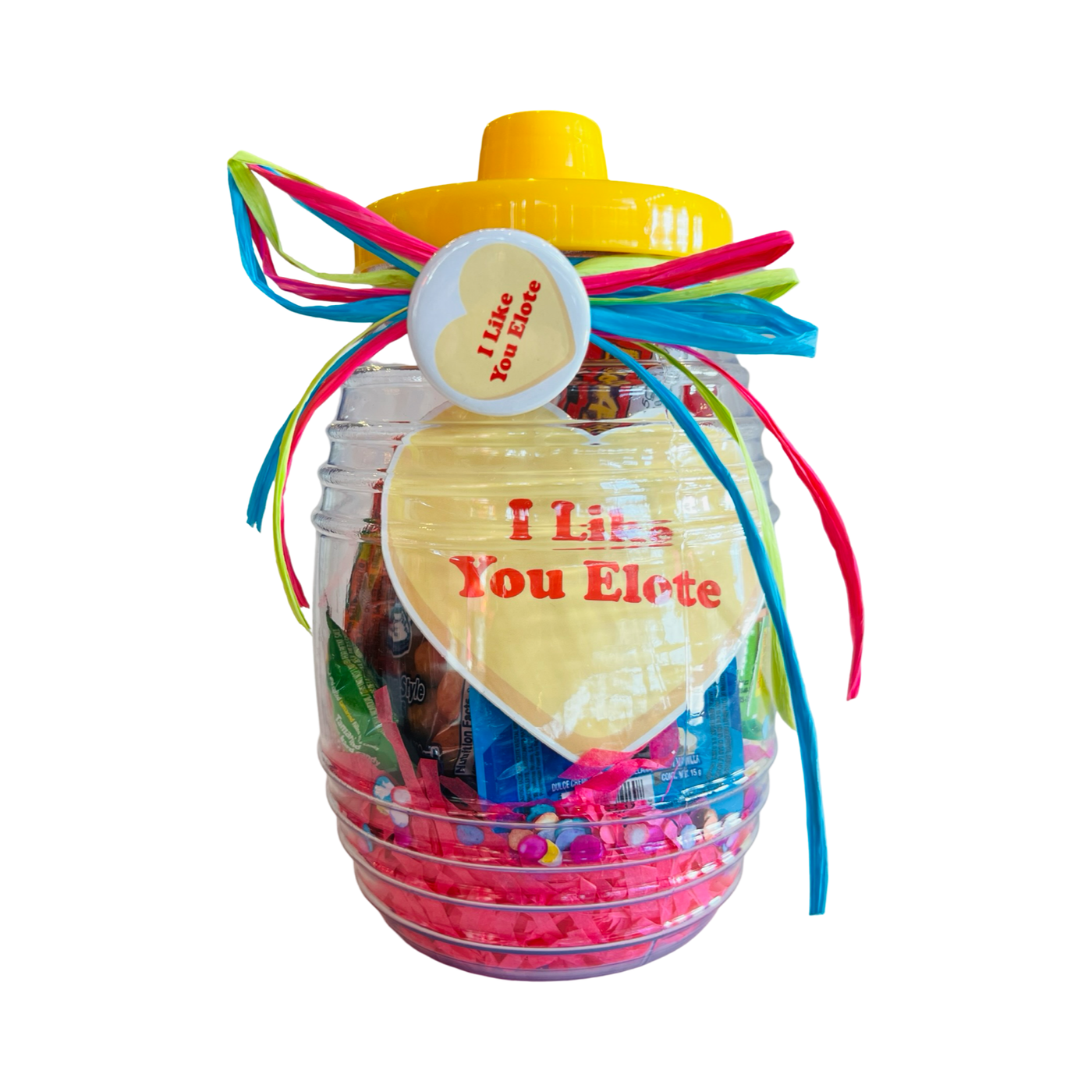 A clear candy jar decorated with vibrant ribbons and filled with colorful confetti, assorted Mexican candies, and a yellow and white pin-back button featuring a yellow heart and I like you elote Phrase design. The jar also includes a heart shaped sticker with the phrase I Like You Elote