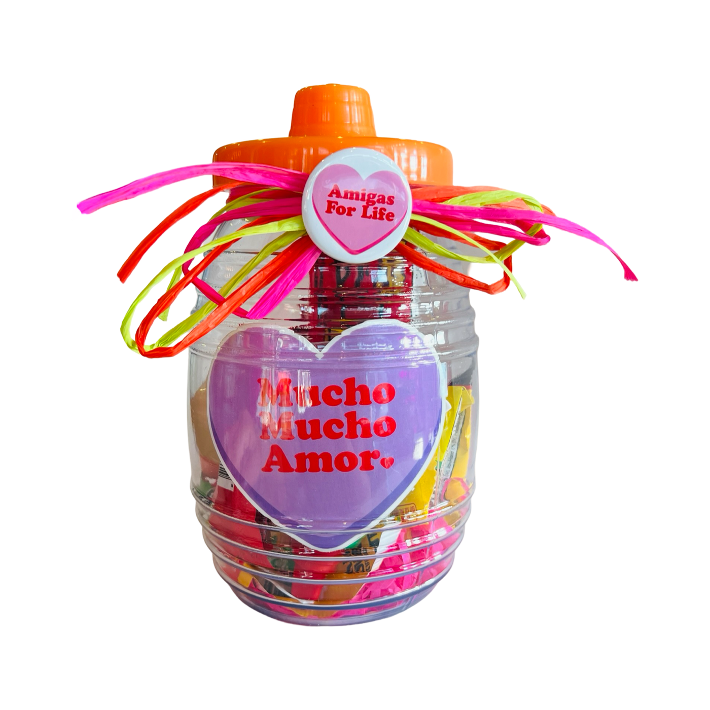 A clear candy jar decorated with vibrant ribbons and filled with colorful confetti, assorted Mexican candies, and a pink and white pin-back button featuring a pink heart and Amigas for Life Phrase design. The jar also includes a heart shaped sticker with the phrase Mucho Mucho Amor