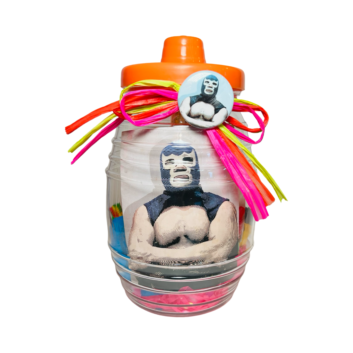 A clear candy jar decorated with vibrant ribbons and filled with colorful confetti, assorted Mexican candies, and a light blue pin-back button featuring a blue masked luchador. The jar also includes a matching luchador sticker