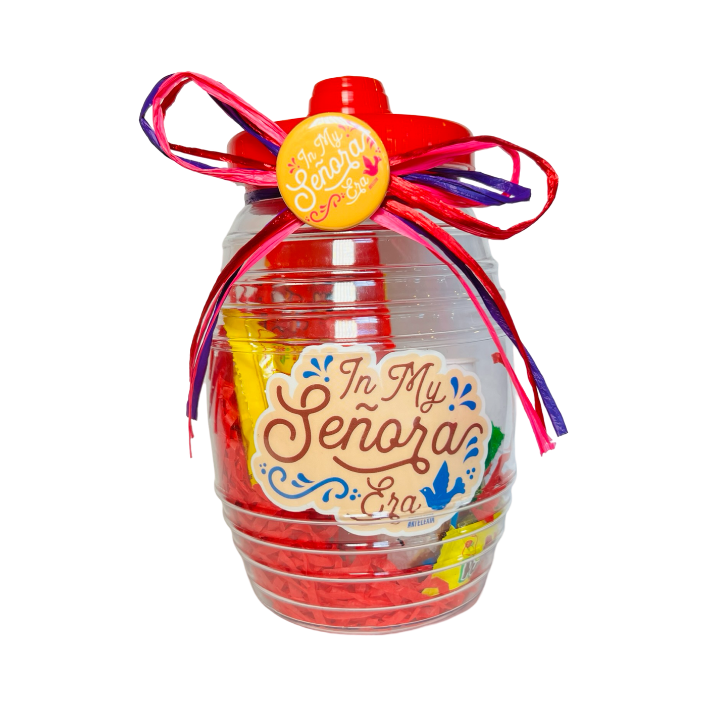 A clear candy jar decorated with vibrant ribbons and filled with colorful confetti, assorted Mexican candies, and a yellow pin-back button featuring the phrase In My Senora Era. The jar also includes a matching sticker.