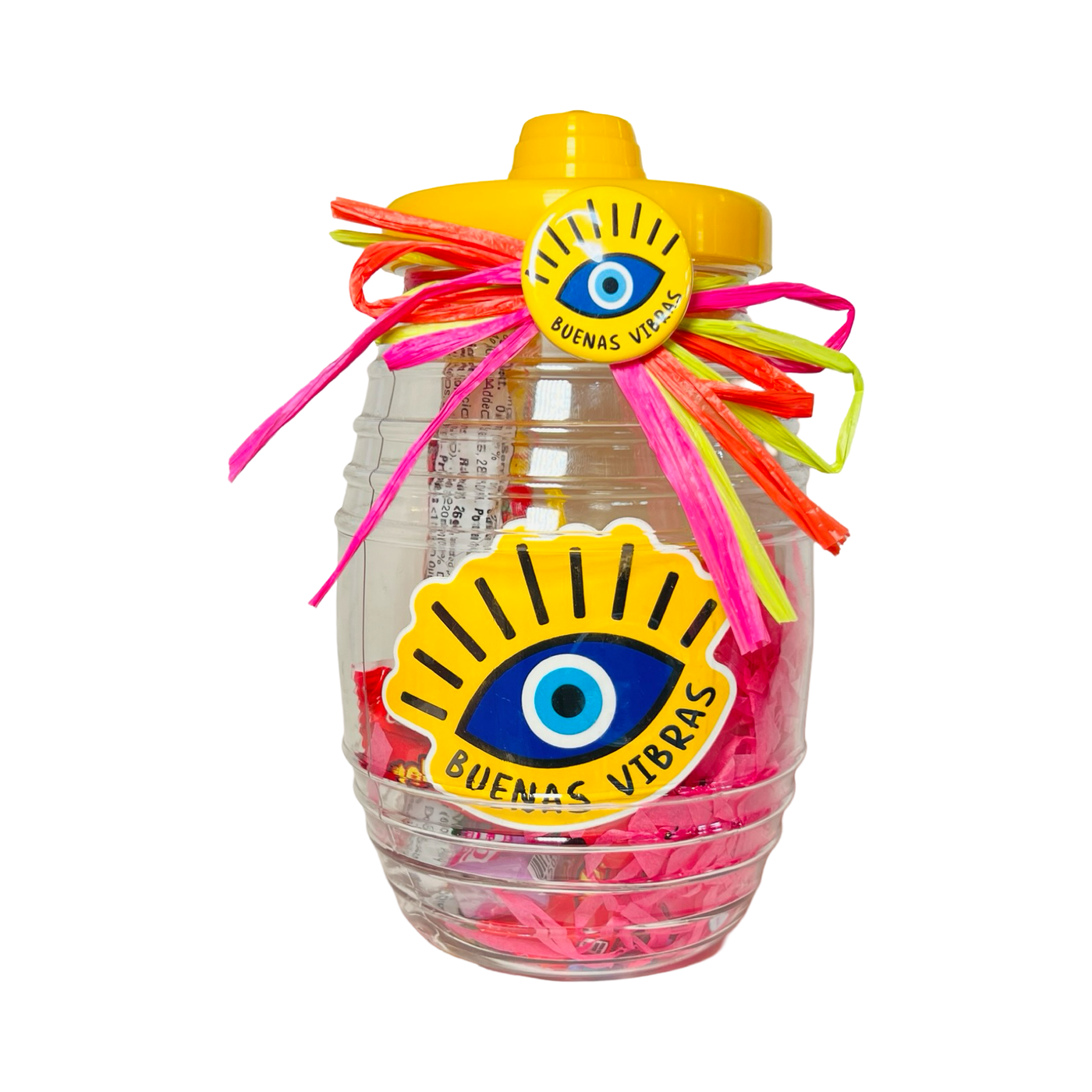 A clear candy jar decorated with vibrant ribbons and filled with colorful confetti, assorted Mexican candies, and a yellow pin-back button featuring an evil eye and the phrase Buenas Vibras. The jar also includes a matching Buenas Vibras sticker.