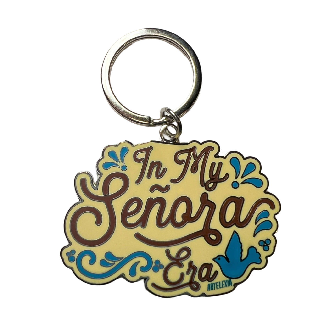 Cream enamel keychain with the phrase In My Senora era in brown lettering and blue filagree and bird.