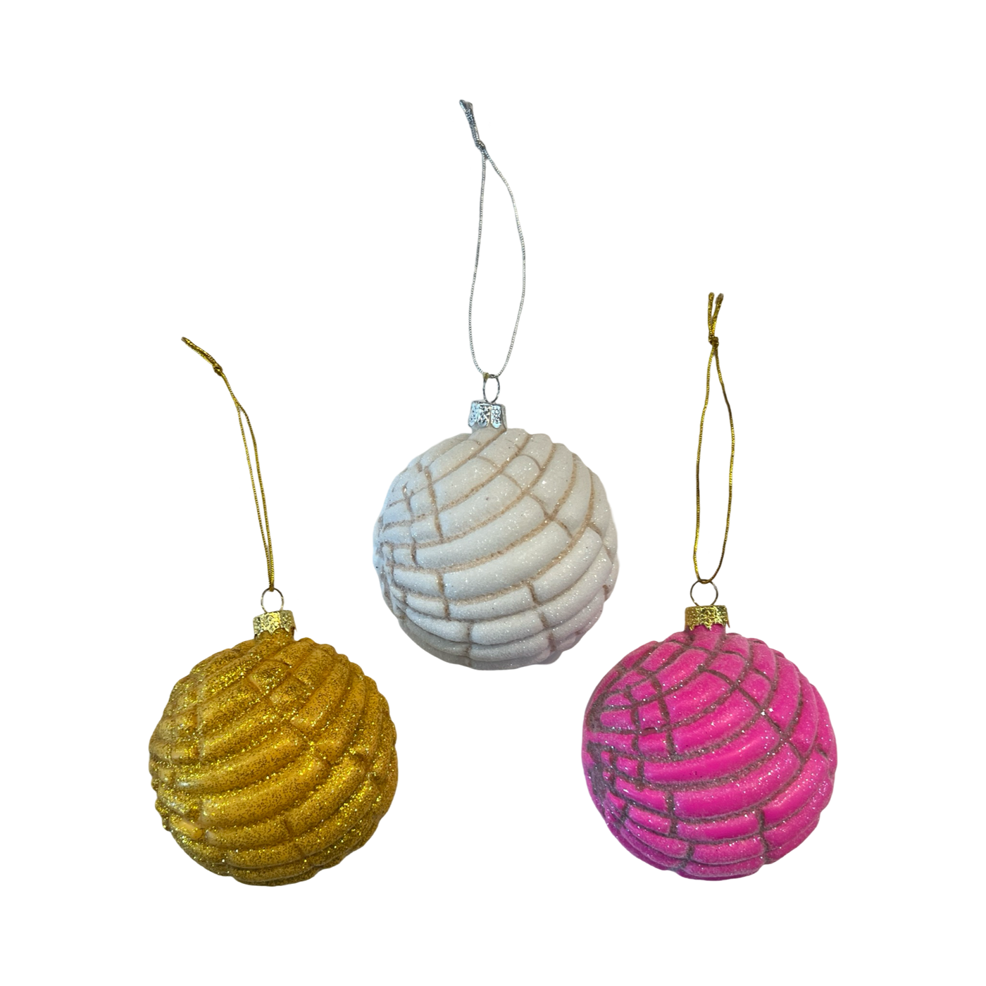 Three glass Christmas ornaments shaped like Mexican conchas, featuring intricate textured designs. The ornaments come in glittery gold, white, and pink, each hanging from a metallic string.