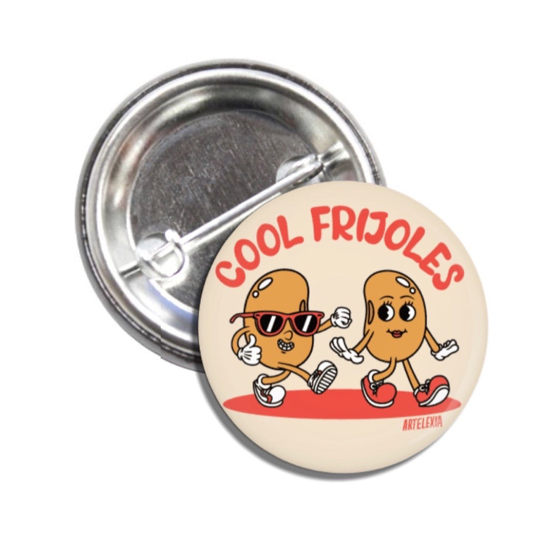 A set of pin-back buttons with one showing the back of the pin and the other on is beige with an illustration of two animated beans and the phrase Cool Frijoles in red lettering