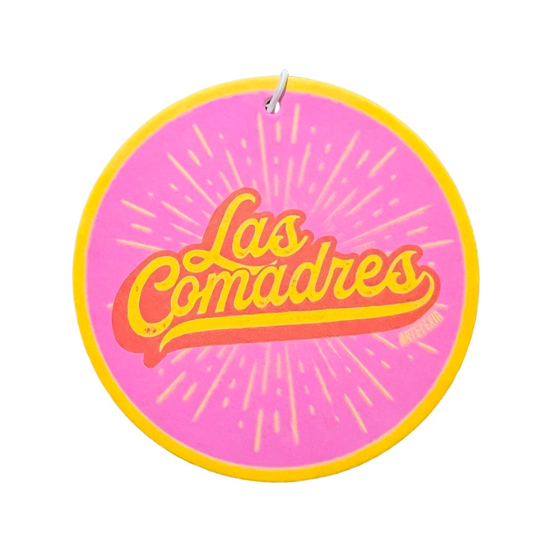 Round pink air freshener with the phrase Las Comadres in yellow and red lettering with a yellow burst in the background.