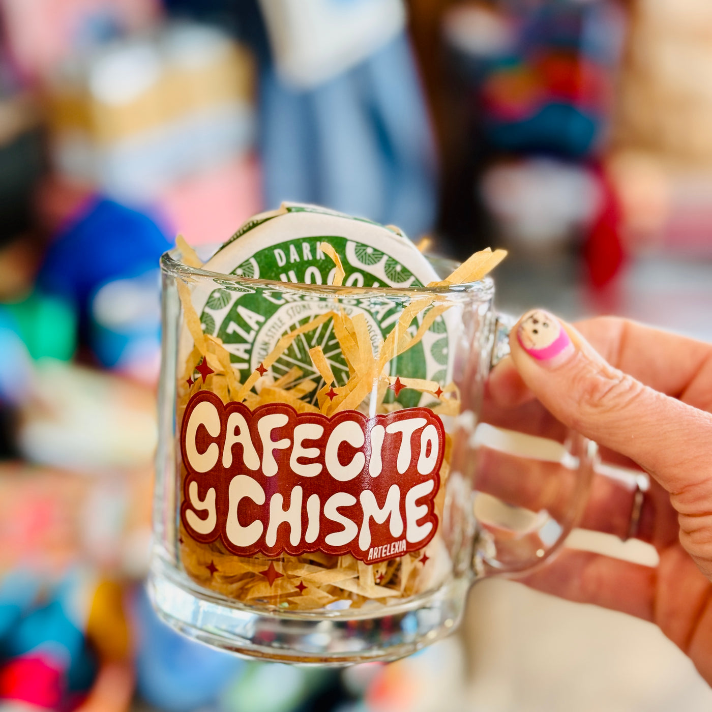 cafecito y chisme clear mug with brown lettering, filled with Kraft crinkle paper and a wrapped Taza chocolate 