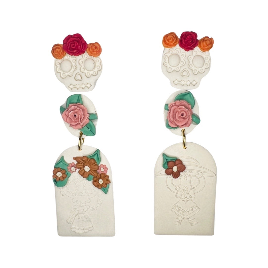 Two sets of clay earrings. One is a set of sugar skulls with red and orange roses on the top. The second set is a dangle style earring with a pink rose and tombstone shaped earring with a calavera design stamped onto it.