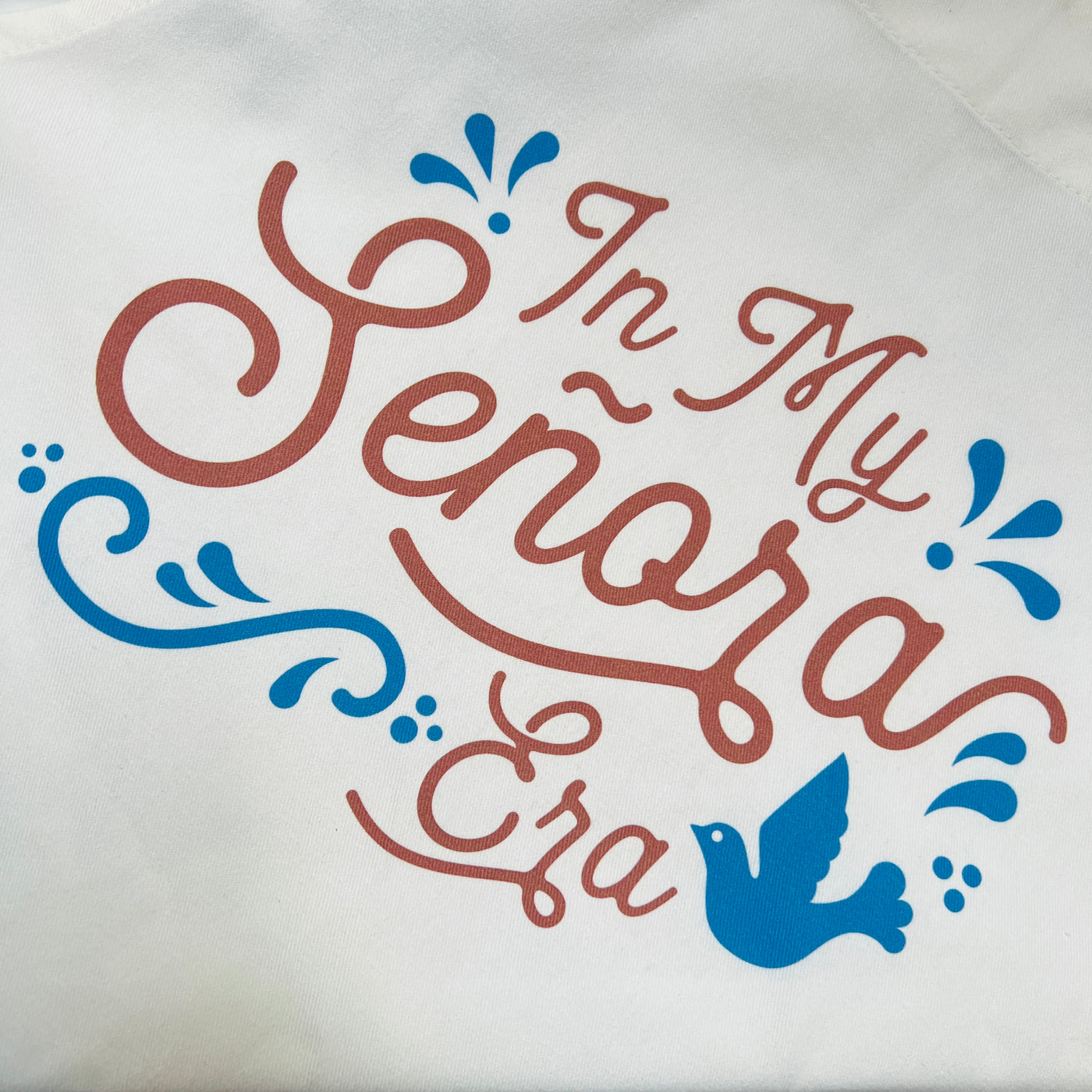 close up view of a white apron with the phrase In My señora era in brown lettering and features a blue bird and filagree