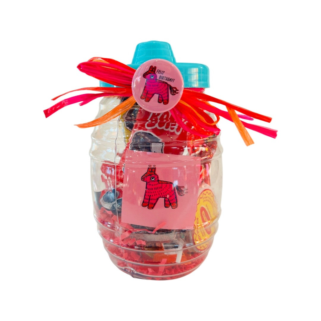 A clear candy jar decorated with vibrant ribbons and filled with colorful confetti, assorted Mexican candies, and a pink pin-back button featuring a piñata design. The jar also includes a matching pink sticker with a playful piñata illustration, bringing festive energy to the design.