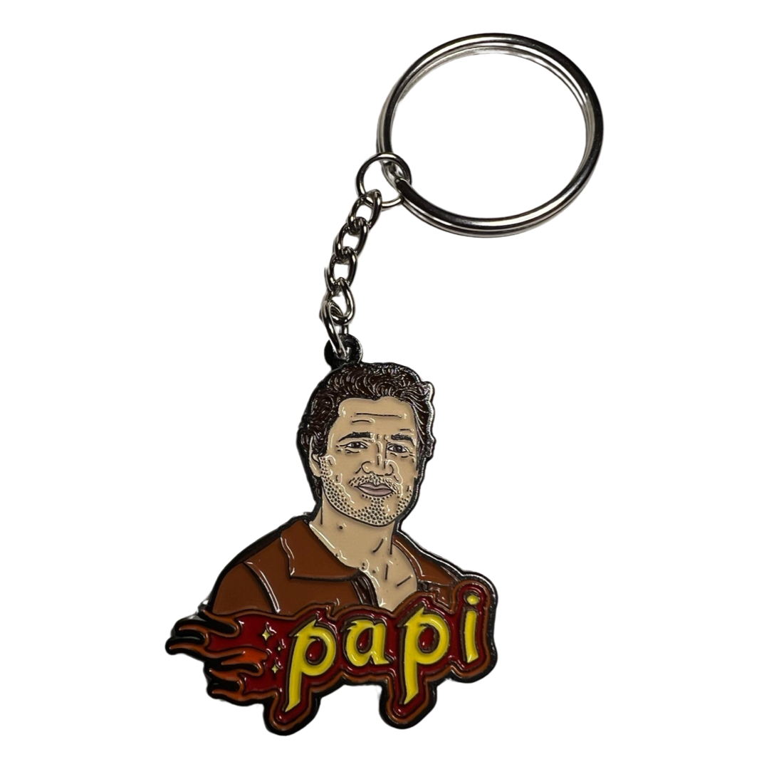Enamel keychain in the shape of Pedro Pascual wearing a brown shirt and the phrase Papi in yellow lettering with some red and orange flames.