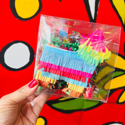 Multi-colored pinata in a clear plastic box with crinkle paper and candy inside. Being held up in front of a red, white and yellow background.