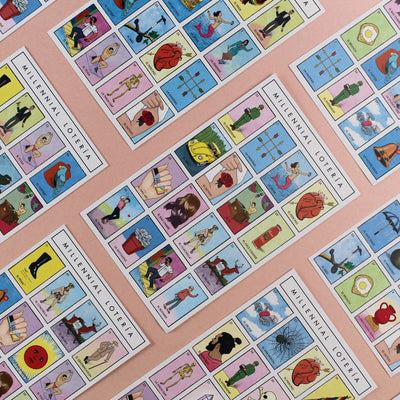 Several colorful Millennial Lotería game boards laid out on a pink background, showing modern illustrated tiles with Spanish labels (e.g., La Selfie, El Corazón).