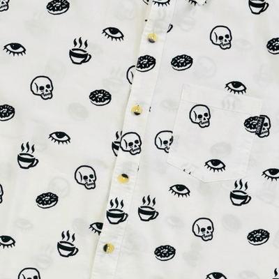 Close up view of a white short-sleeve button-down shirt with allover black prints of coffee cups, donuts, skulls, and eyes.