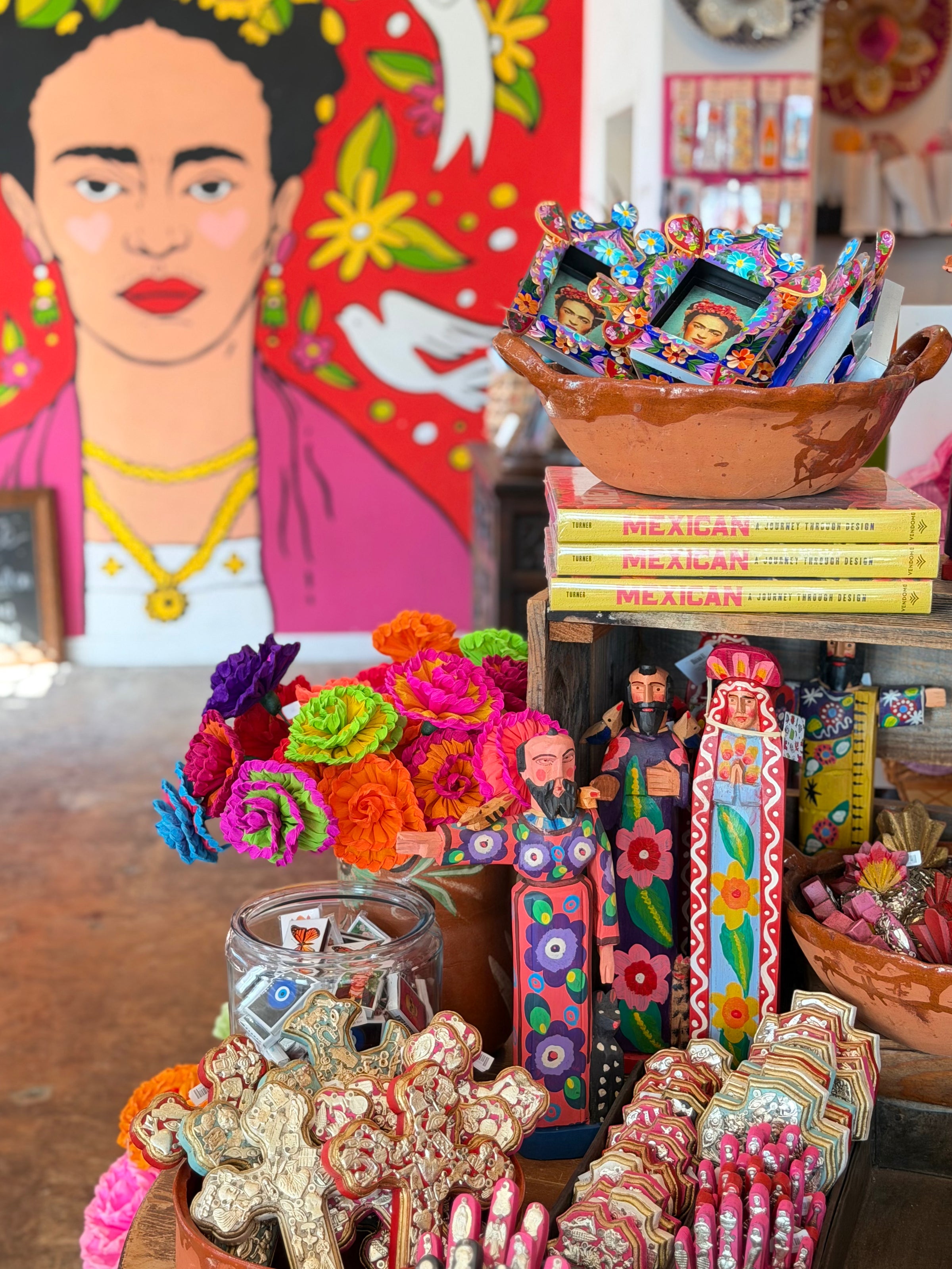 A colorful artisanal display in a shop featuring hand-painted wooden figurines, bright marigold flowers, patterned cookies, and packaged snacks stacked on books in front of a large Frida Kahlo-style portrait painting.