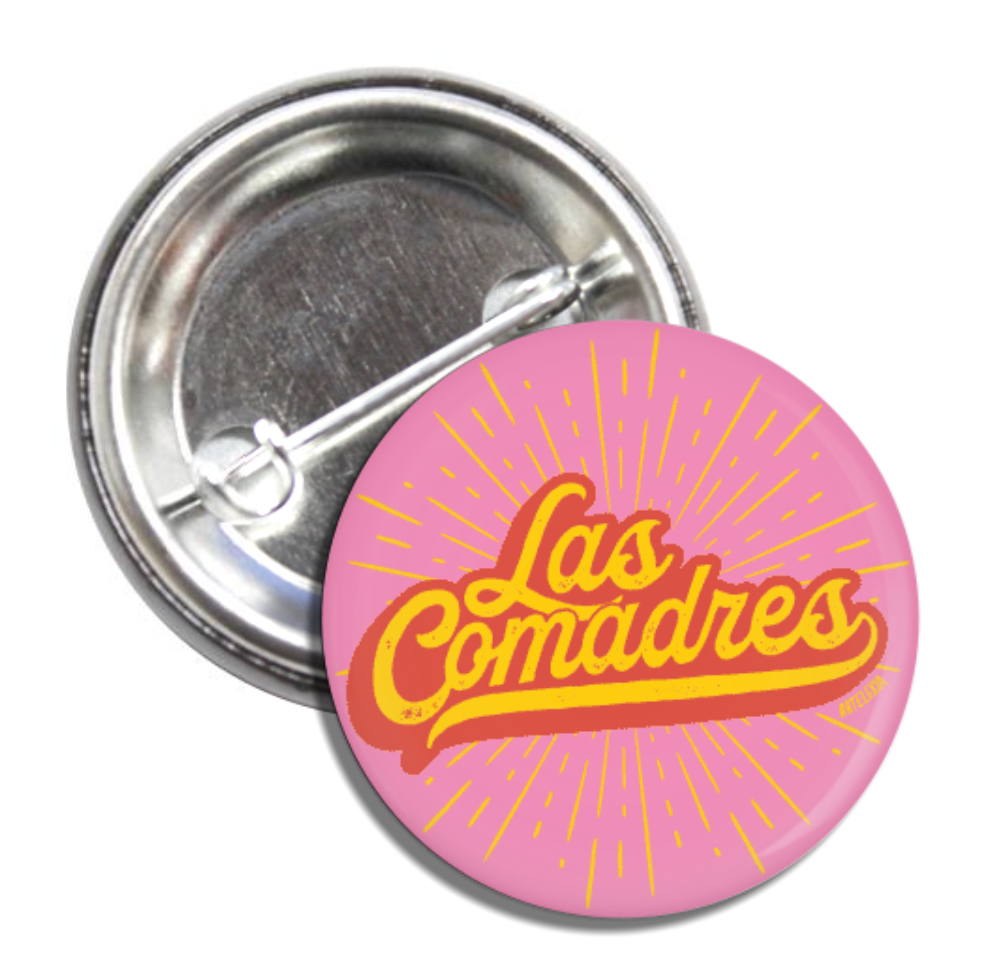 Round pin back button with a pink background with the phrase Las Comadres in yellow and orange lettering.