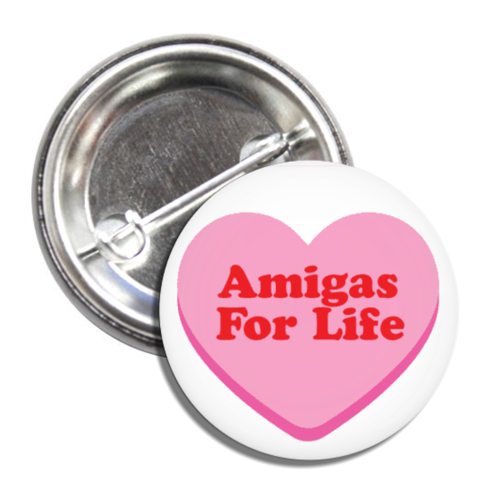 Round white pin back button featuring a pink heart and the phrase Amigas For Life in red lettering.