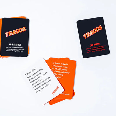 Tragos game cards spread out on a white surface, featuring a mix of black, orange, and white cards with bilingual prompts in English and Spanish, including playful instructions and challenges.