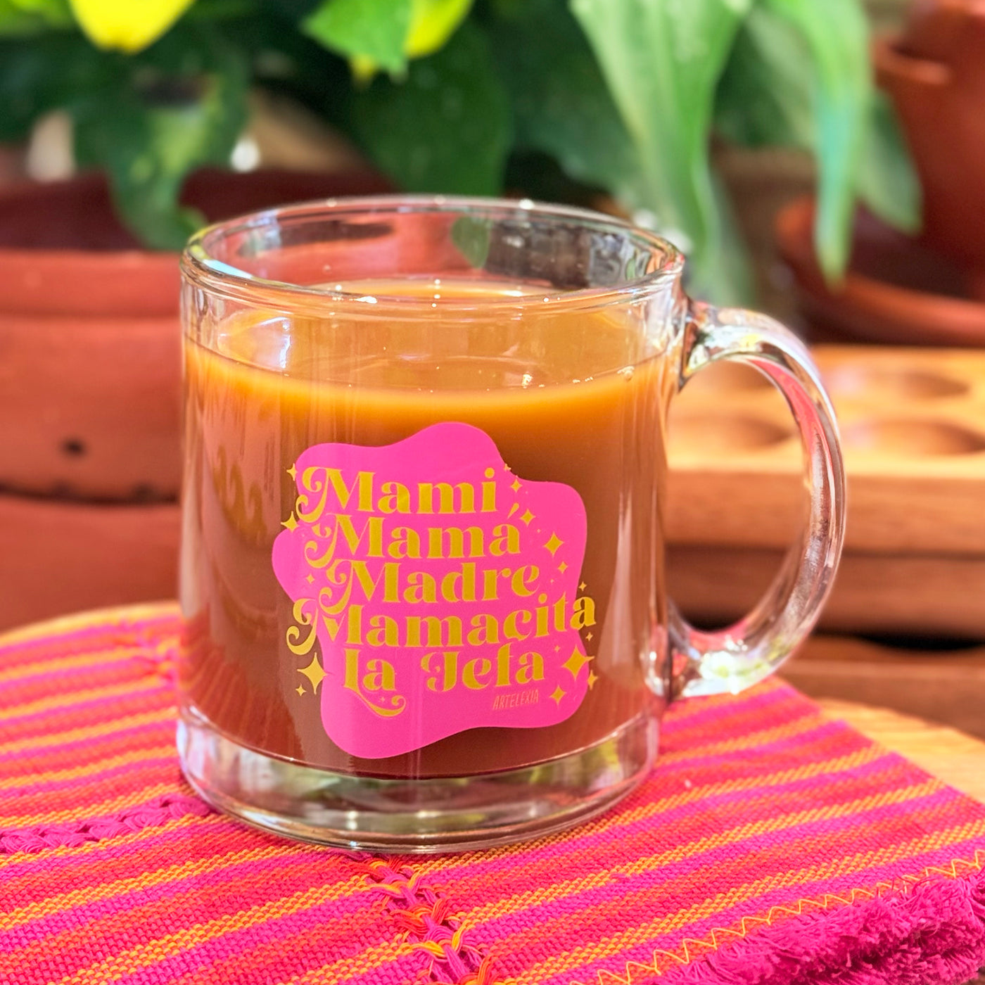 Photo of clear glass mug with phrase "Mami, Mama, Madre, Mamacita, La Jefa" in gold lettering on top of a magenta shape