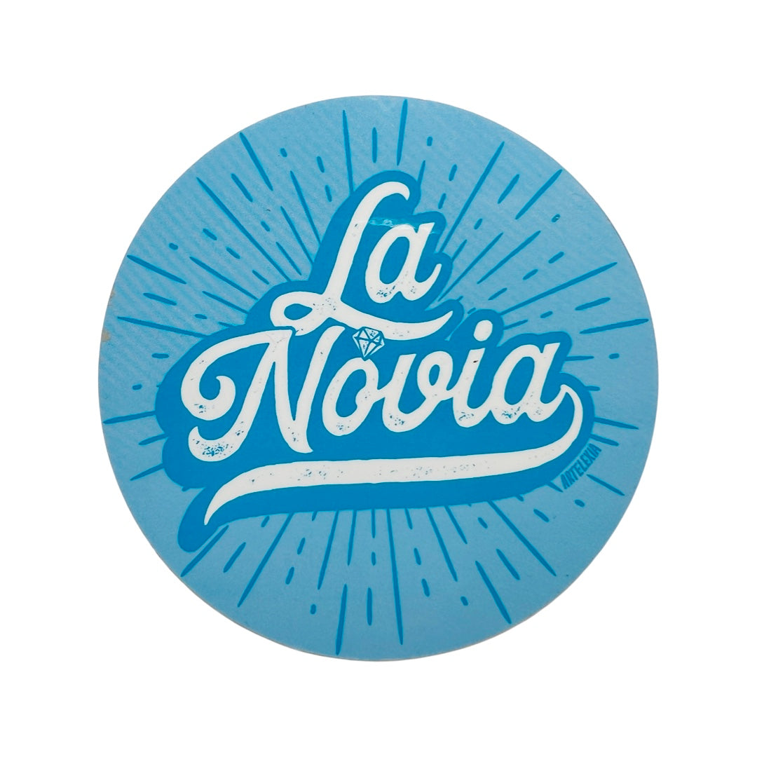 Round light blue circle sticker with the phrase La Novia in white lettering and features a diamond above the lettering "O."