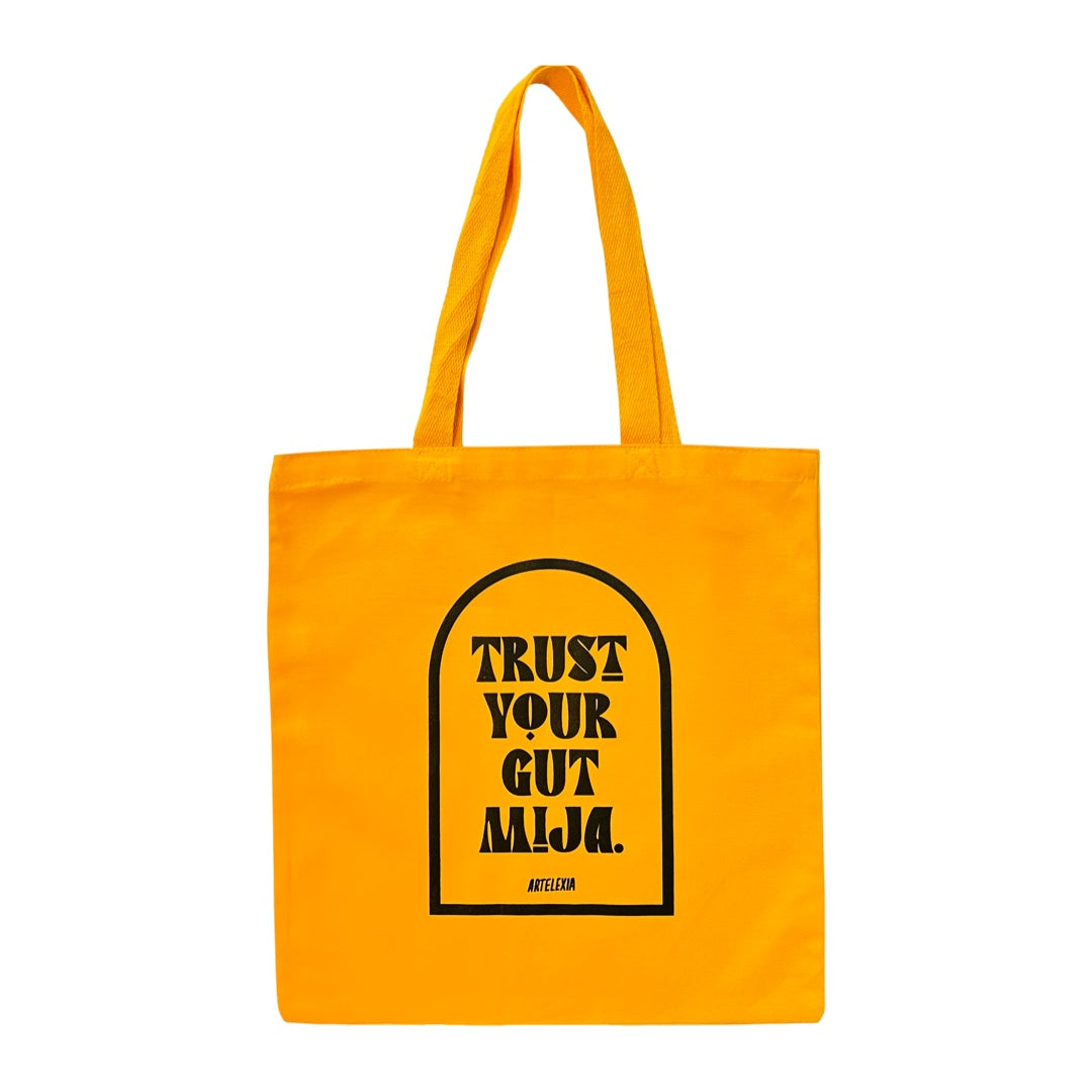 Yellow canvas tote bag with the phrase Trust Your Gut Mija in black lettering with a black outline of an arch surrounding it.