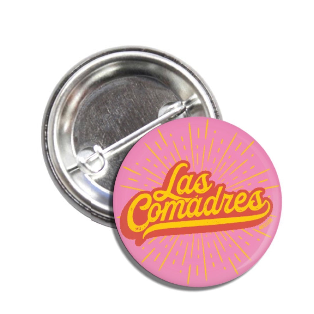 Set of pin-back buttons with one shoing the back of one and the other one is pink with a yellow burst and the phrase Las Comadres in red and yellow lettering.