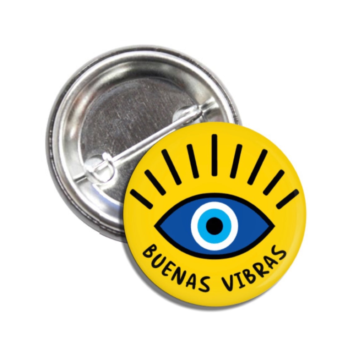 Set of pin-back buttons with one showing the back of it and the other one is yellow with an illustration of an eye and the phrase Buenas Vibras in black lettering.