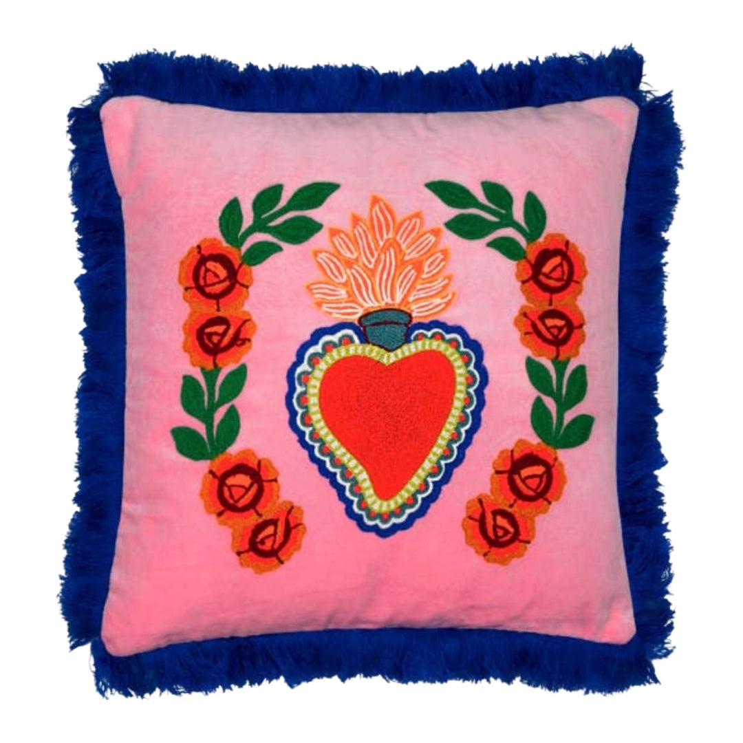 Square pink pillow with a central embroidered red and blue heart, floral accents, and bold blue fringe trim.