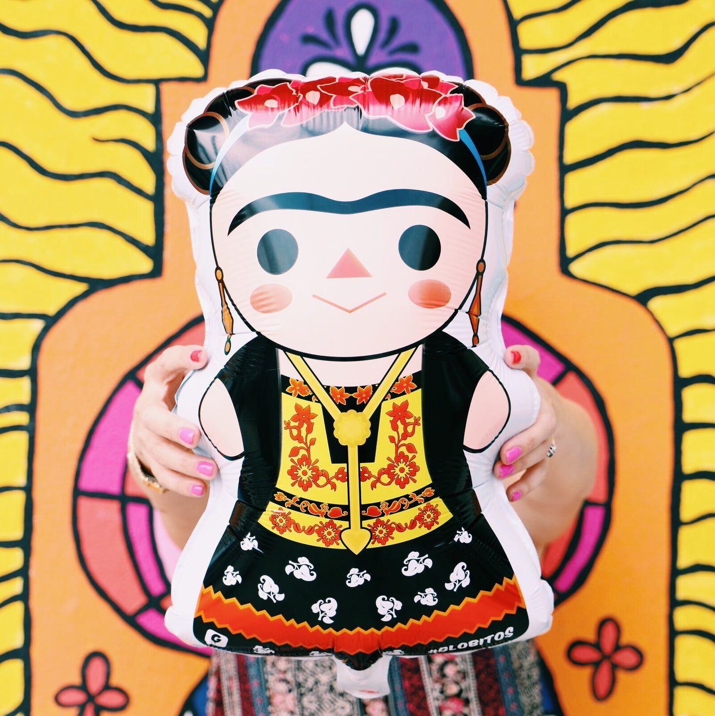 Frida Kahlo balloon photographed in front of Artelexia's, "You Are Radiant" mural. Frida has a Maria doll style appearance. Main colors featured are red, black, and yellow. Frida wears a red flower crown and traditional dress.