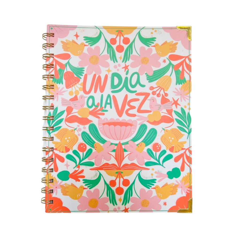 Hardcover notebook that features a design of various plants, cacti and birds in an orange, green and pink color palette. In the center the phrase Un Dia A La Vez. Translation: one day at a time